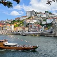 Traditional wooden boat with passengers on the Douro River in Porto, with colorful hillside buildings and the iconic Dom Luís I Bridge visible.