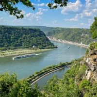 Panoramablick auf den Rhein, der sich durch grünes Tal windet, mit Flusskreuzfahrtschiff, bewaldeten Hügeln und Stadt am Ufer vom Loreleyfelsen.