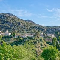 Weiße Dorfhäuser der Alpujarras zwischen grünen Hügeln und Bergen unter blauem Himmel, mit markanter Felsspitze im Hintergrund.