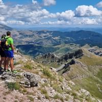 Two hikers with backpacks standing on rocky summit of Bjelasnica mountain in Bosnia, overlooking vast mountain landscape with valleys below.
