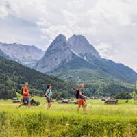 Drei Wanderer mit Rucksäcken gehen durch grüne Wiesen bei Garmisch mit dramatischen Alpengipfeln im Hintergrund unter bewölktem Himmel.