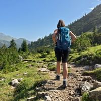 Wanderer mit blauem Rucksack auf felsigem Bergweg durch grünes Alpental mit bewaldeten Hängen und Berggipfeln unter blauem Himmel.