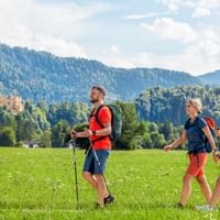 Three hikers with backpacks walking across a green meadow near Hohenschwangau. Forested mountains and a castle are visible in the background.