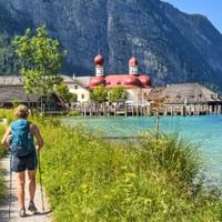 Wanderin mit Rucksack auf Uferweg am Königssee. St. Bartholomä Kirche mit roten Kuppeln über türkisfarbenem Wasser, Berge im Hintergrund.