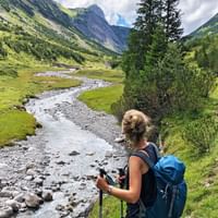 Female hiker with blue backpack and trekking poles on Lechweg, overlooking a green mountain valley with a winding stream.