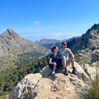 Two hikers sitting on rocky outcrop on GR221 trail in Mallorca, overlooking mountain valley with Puig Major peak and blue sky.