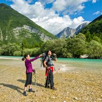 Wanderpaar mit Fernglas am steinigen Ufer der türkisen Soca, mit den Bergen im Hintergrund