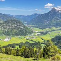 Alpentalblick von der Buchensteinwand bei Kitzbühel mit grünen Wiesen, bewaldeten Hügeln, kleinen Dörfern und schneebedeckten Bergen unter blauem Himmel.