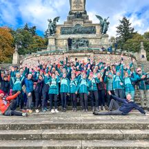 Menschengruppe mit blauen Jacken heben die Hände in die Höhe, stehen vor einem Monument