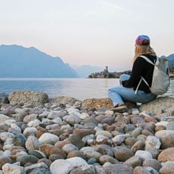 Woman with backpack sitting on rocky shore of Lake Garda, viewing mountains and Malcesine town across calm water at dusk.