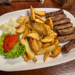 White plate with grilled ćevapčići, golden fries, sliced onions, red ajvar sauce on lettuce, served on wooden table in Soča Valley.