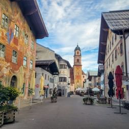 Fußgängerzone in Mittenwald mit bunten Lüftlmalerei-Häusern und barockem Kirchturm. Pflanzkübel und Außengastronomie säumen die Straße unter blauem Himmel.