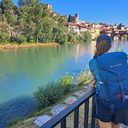 Person with blue backpack viewing historic covered bridge and colorful buildings of Bassano del Grappa from riverside path along green river.