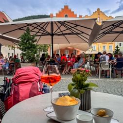 Rosa Rucksack neben Tisch mit Getränk, Chips und Blumen in Café in Sterzing. Bunte historische Gebäude und Sonnenschirme im Hintergrund.