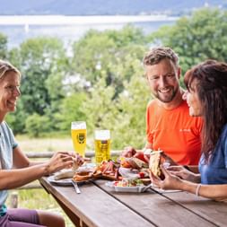 Drei Wanderer teilen traditionelle bayerische Brotzeit mit Brot, Aufschnitt und Bier an Holztisch im Freien mit grünen Bäumen im Hintergrund.