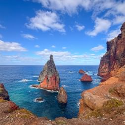 Dramatic coastal view at São Lourenço, Madeira with pointed rock formations rising from blue ocean waters, surrounded by steep red-brown cliffs.