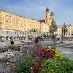 Colorful flower beds on Hauptplatz square in Linz with yellow historic buildings and twin-towered church in the background under blue sky.