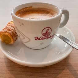 White cup of cappuccino with foam on saucer, accompanied by a cannolo pastry dusted with powdered sugar and a spoon.