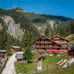 Traditionelle Berndlalm Holzhütte mit roten Fensterläden im Obersulzbachtal, umgeben von grünen Wiesen, Wald und felsigen Gipfeln unter blauem Himmel.