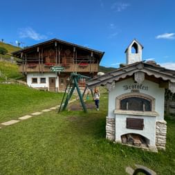 Traditional Alpine farmhouse with wooden balcony and white chapel tower, playground, and historic bread oven labeled 'Brotofen' on green meadow.