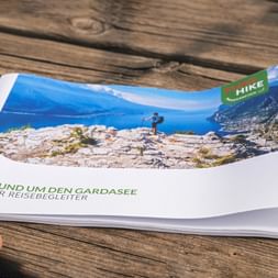 Hand holding a travel brochure titled 'Rund um den Gardasee' showing a hiker on rocky terrain overlooking Lake Garda's blue waters.