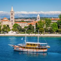MS Dalmatino Holzsegelschiff mit gelber Kabine im türkisfarbenen Hafenwasser. Historische Kirchtürme und Uferpromenade sichtbar.