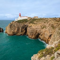 Leuchtturm Cabo de São Vicente mit roter Spitze auf dramatischer Klippe mit Blick auf türkisfarbenen Atlantik und Felsformationen.