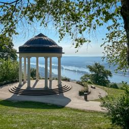 White columned pavilion with dark dome on a hilltop overlooking the Rhine River valley. Green trees frame the view under a blue sky.