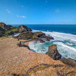 Felsige Klippen an der Praia da Pena Furada mit türkisfarbenen Meereswellen, die gegen Felsformationen unter blauem Himmel schlagen.