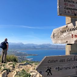 Wanderer am Mallorca Bergaussichtspunkt Wanderer mit Rucksack steht an hölzernen Wegschildern mit Blick auf Mallorcas Küste und Berge unter blauem Himmel.