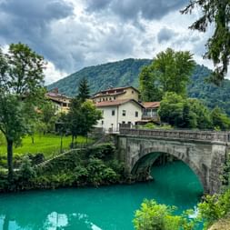 Historic stone arch bridge spanning turquoise Soča River near Tolmin. White buildings and forested mountains of the Julian Alps under cloudy sky.