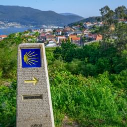 Steinmarkierung mit gelber Jakobsmuschel auf blauem Grund am Portugiesischen Jakobsweg bei Vigo mit Blick auf Küstenstadt und Bucht.
