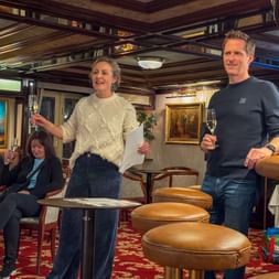 Group celebration on elegant ship interior with wooden ceiling, red carpet. Woman and man holding champagne glasses, others seated in background.