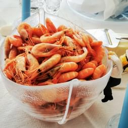 Große Glasschale gefüllt mit gekochten orangefarbenen Garnelen auf weißer Tischdecke, umgeben von blauen Kerzen, Gläsern und weißen Tellern.