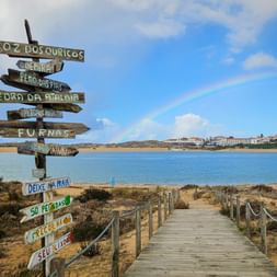 Hölzerner Wegweiser mit mehreren Richtungspfeilen am Rota Vicentina Wanderweg. Regenbogen über Rio Mira mit Vila Nova de Milfontes im Hintergrund.