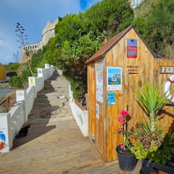 Hölzerne Fährstation mit Rettungsring auf Holzsteg in Vila Nova de Milfontes, Alentejo. Weißes Geländer und Steinfestung auf Hügel sichtbar.