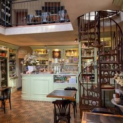 Two-story delicatessen interior with spiral staircase, mint-green shelving, wooden tables, and products displayed on walls and counters.