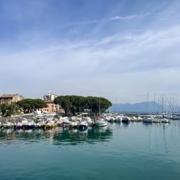 Hafen von Desenzano del Garda mit Booten und Yachten im türkisfarbenen Wasser. Historische Gebäude und Bäume säumen das Ufer, Berge im Hintergrund.
