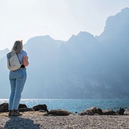 Woman with backpack standing on rocky shore of Lake Garda, facing dramatic mountain peaks across the blue water under hazy sky.