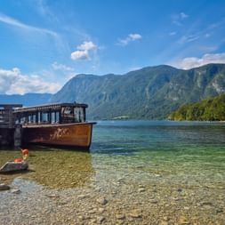 Wooden excursion boat moored at a pier on Lake Bohinj with clear shallow water, mountains in the background, and blue sky with clouds.