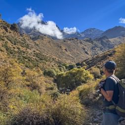 Wanderer mit Rucksack betrachtet ein Bergtal in den Alpujarras mit herbstlicher Vegetation, felsigen Hängen und weißen Wolken über Gipfeln.