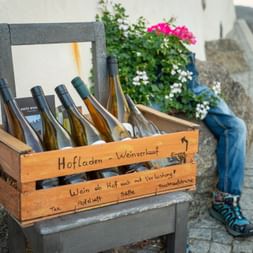 Holzkiste mit Weinflaschen auf Stuhl in Partschins, Südtirol. Schild mit 'Hofladen-Weinverkauf', daneben sichtbare Beine einer Person.