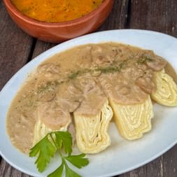White plate with rolled pasta topped with creamy mushroom sauce and herbs, served on a wooden table. A terracotta bowl with soup is visible above.