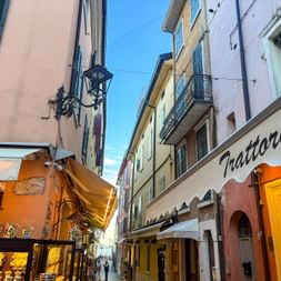 Enge Gasse in Garda mit bunten Gebäuden und einer Trattoria. Gelbe Markisen und Geschäfte säumen die Fußgängerzone.