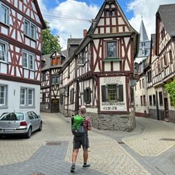 Wanderer mit Rucksack auf dem Marktplatz von Braubach, umgeben von traditionellen Fachwerkhäusern und einem Kirchturm im Hintergrund.
