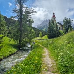 Wanderweg neben einem Bach zur Nikolauskirche in Obernberg am Brenner, mit grünen Wiesen, Kiefern und Bergen.