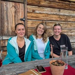 Three smiling women sitting at wooden table outside traditional alpine hut. They wear casual outdoor clothing and have drinks on the table.