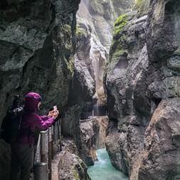 Wanderin in lila Jacke fotografiert mit Handy in der Partnachklamm nahe der Zugspitze. Enge Felsschlucht mit türkisfarbenem Bach.