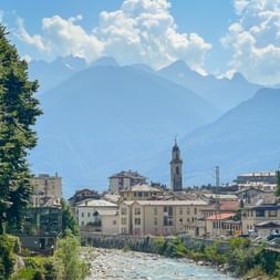 View of Chiavenna with historic buildings and church tower along a river, framed by trees. Layered mountain peaks rise under a cloudy sky.