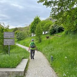 Wanderer mit grünem Rucksack auf gepflastertem Weg zum Kloster Eberbach. Schild zeigt 'Klosterschänke Pfortenhaus' am grünen Hang.